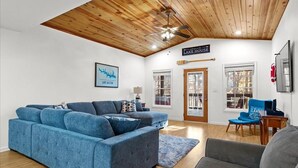 Un salon moderne avec un canapé d’angle bleu, un fauteuil gris et un plafond en bois. Une inscription sur le mur indique : « MAISON AU BORD DU LAC ».