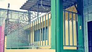 Un edificio con fachada verde y amarilla, una cerca de metal y una ventana con barrotes.