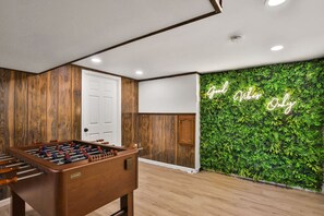 A room with a wooden floor, a foosball table, a white door, and a green wall with a neon sign.