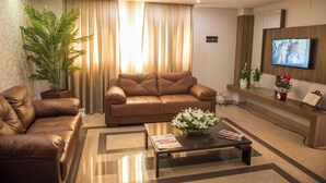 A modern living room with a brown leather sofa, a coffee table with magazines, a large potted plant, and a television on the wall. - LARANJEIRAS PALACE HOTEL (Laranjeiras do Sul)