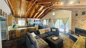 A cozy cabin interior with a large sofa, ottoman, wooden ceiling, kitchen area, and a fireplace. - The cozy cabin! (Twain Harte)