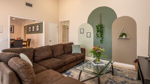 A living room with a brown sectional sofa, a glass coffee table with a vase, and a green wall with decorative arches. - Huge Backyard, Near Disney, Family Friendly, Quiet Neighborhood (Placentia)