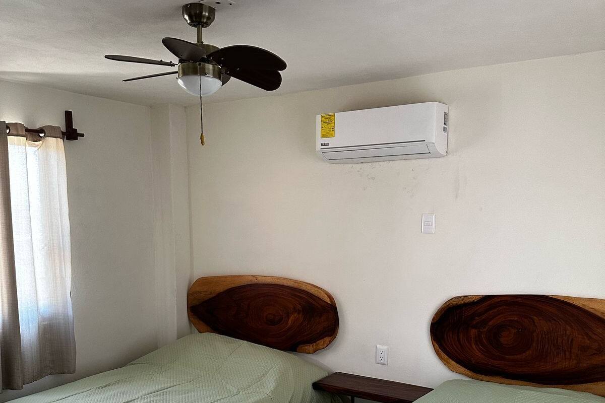 A room with two beds, a ceiling fan, an air conditioner, and a window with curtains.