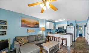 Uma sala de estar com sofá, puf e mesa de centro, uma cozinha com geladeira, micro-ondas e fogão, e um ventilador de teto.