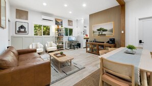 Un salon moderne avec un canapé en cuir marron, une table basse en bois et un coin repas comprenant une table et des chaises blanches. On y trouve également une bibliothèque remplie de divers objets et un téléviseur posé sur un meuble.