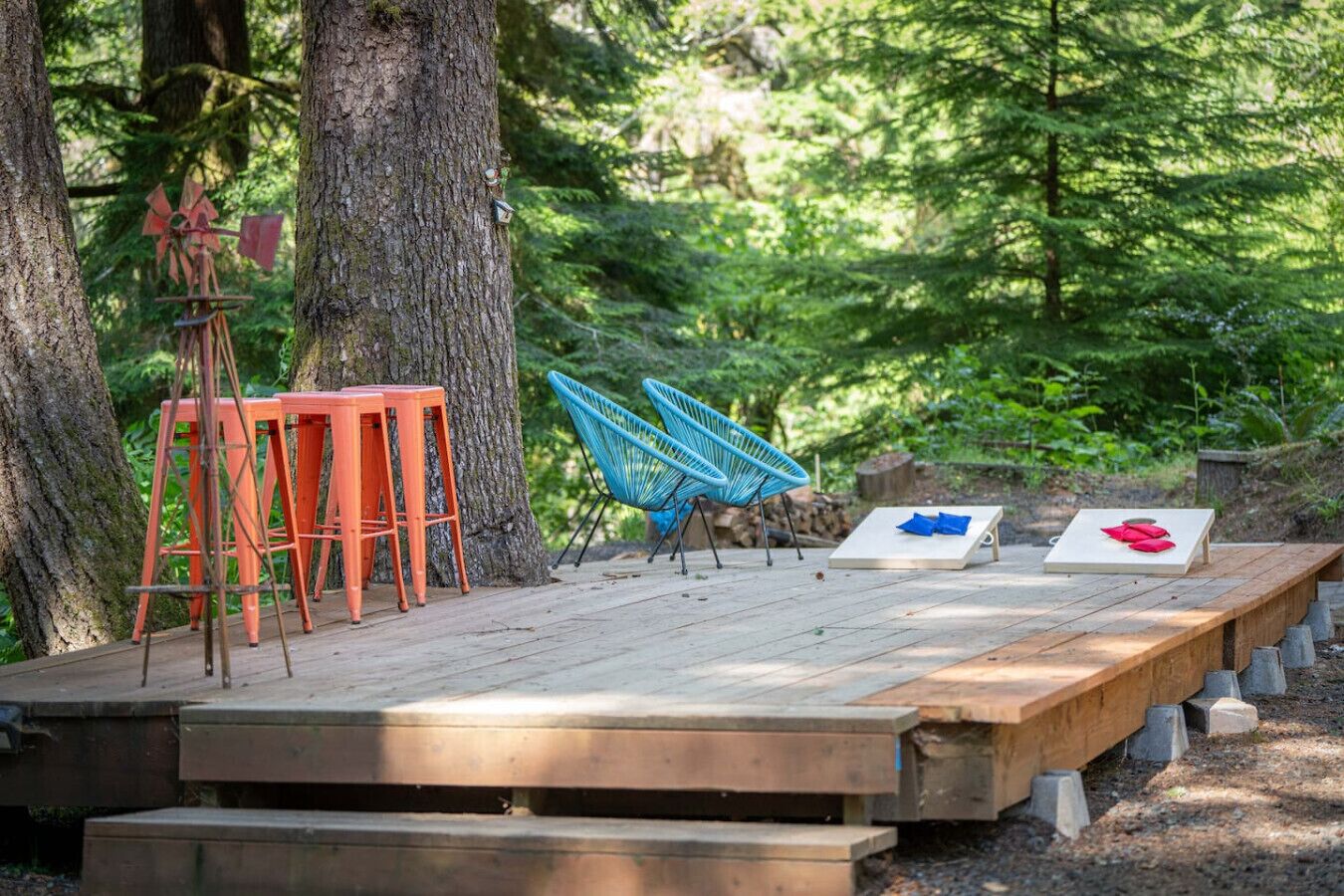 Une terrasse en bois avec des sièges d’extérieur, comprenant des tabourets orange et des chaises bleues, le tout entouré d’arbres.