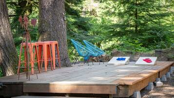 Une terrasse en bois avec des sièges d’extérieur, comprenant des tabourets orange et des chaises bleues, le tout entouré d’arbres.