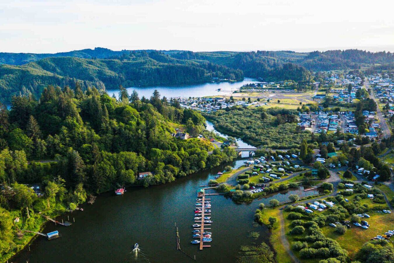 Vue aérienne d’une ville au bord d’un lac, avec un port de plaisance, des collines densément boisées et un petit pont reliant la ville au lac.