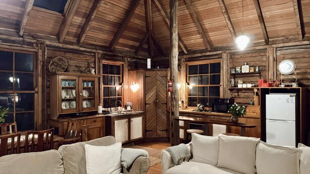 L’intérieur d’un chalet rustique est doté d’un plafond en bois, d’un coin repas avec une table et des chaises en bois, d’une cuisine équipée d’un réfrigérateur et d’étagères, et d’un coin salon confortable avec des coussins.