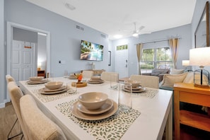 A dining area with a long table set for four, a television mounted on the wall, and a window with curtains.