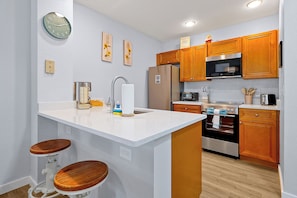 A kitchen with wooden cabinets, a stainless steel refrigerator, a microwave, and a sink with a faucet. There are two stools and a clock on the wall.