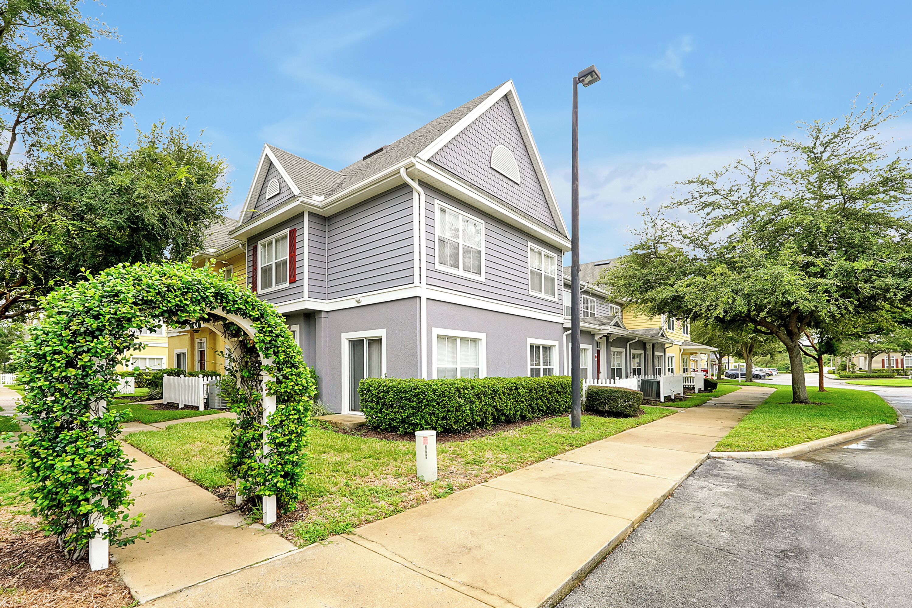 A two-story house with a grey exterior and white trim, a front yard with a hedge, and a sidewalk leading to the street.