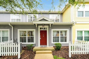 A two-story house with a red door, white picket fence, and a welcome mat at the entrance.