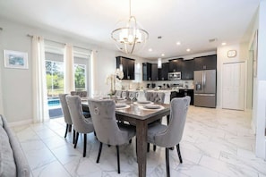 A modern kitchen with dark cabinets, stainless steel appliances, and a dining area with a marble floor and grey upholstered chairs. - 6-Bedroom Retreat in Champions Gate (Four Corners)
