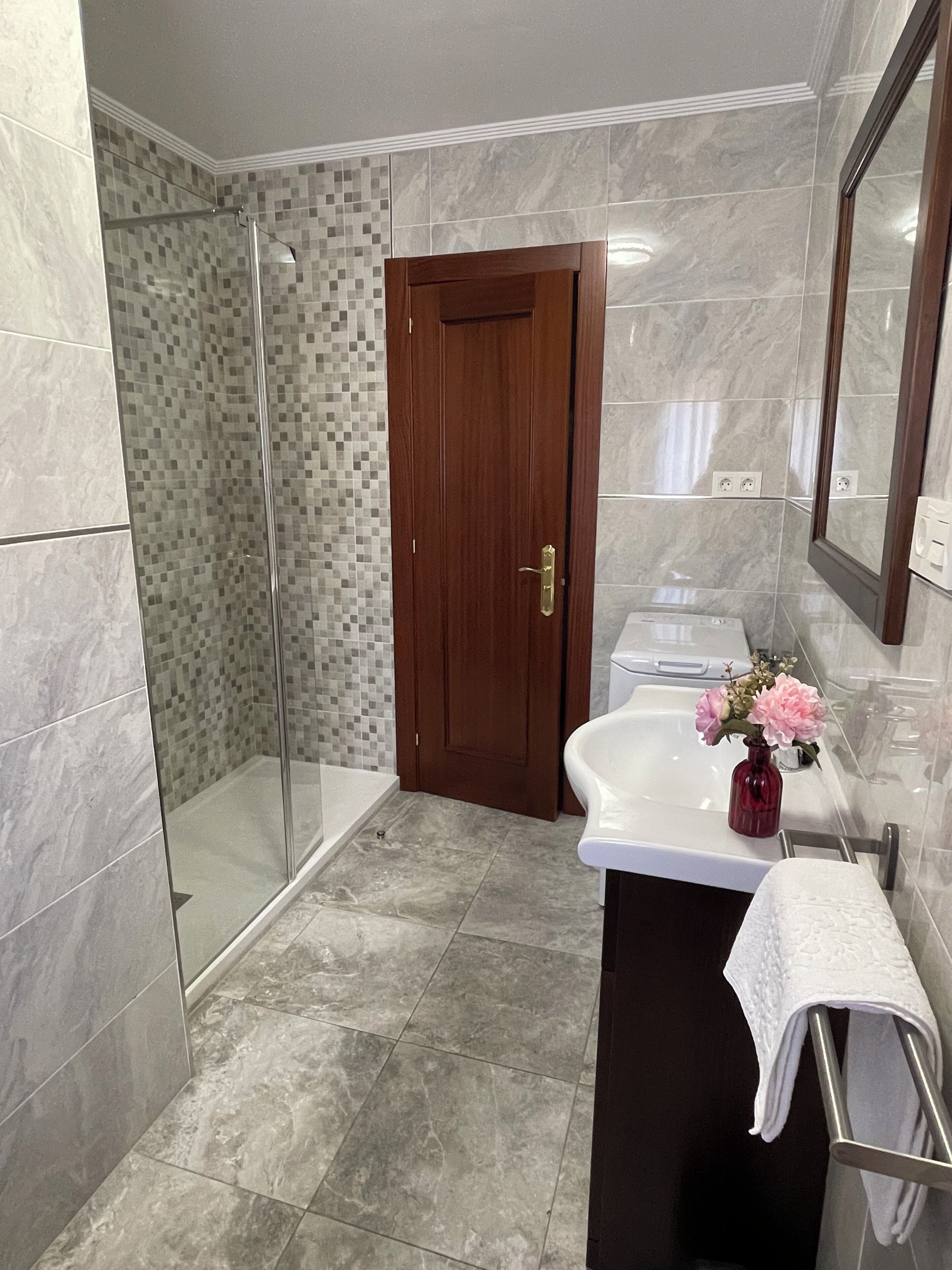 A modern bathroom with a glass shower enclosure, a wooden door, a white sink with a vase of flowers, and a large mirror.