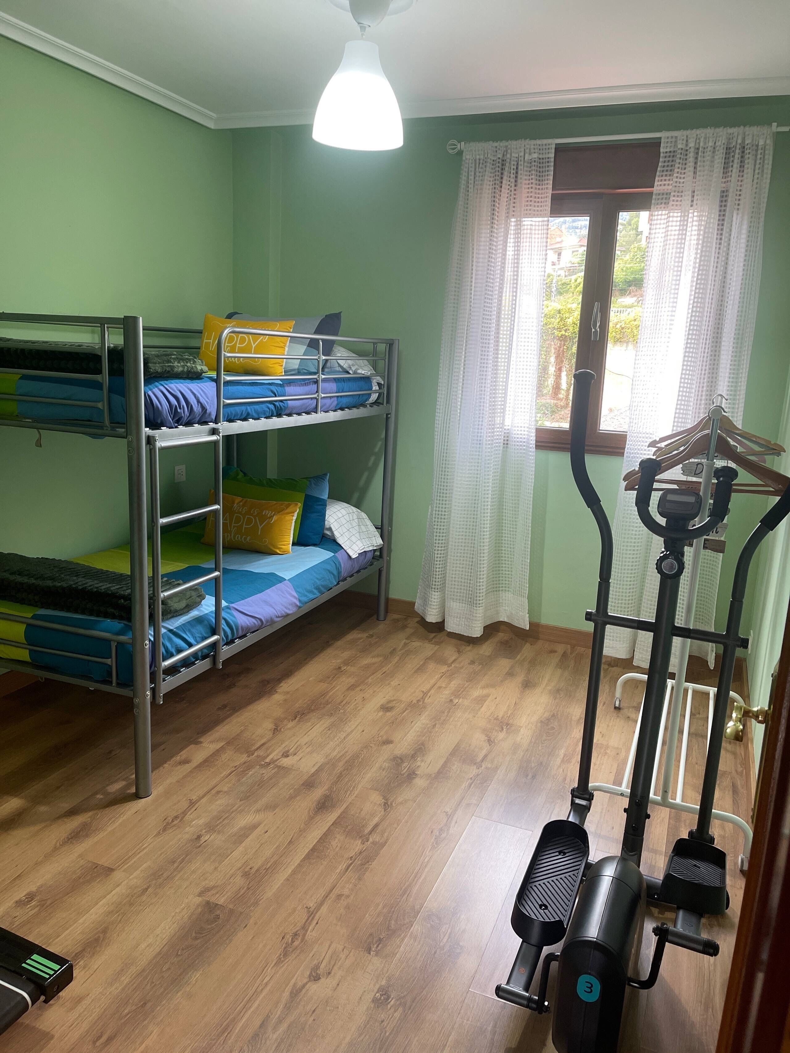 Un dormitorio con literas, ventilador de techo, ventana con cortinas y una máquina de ejercicios.