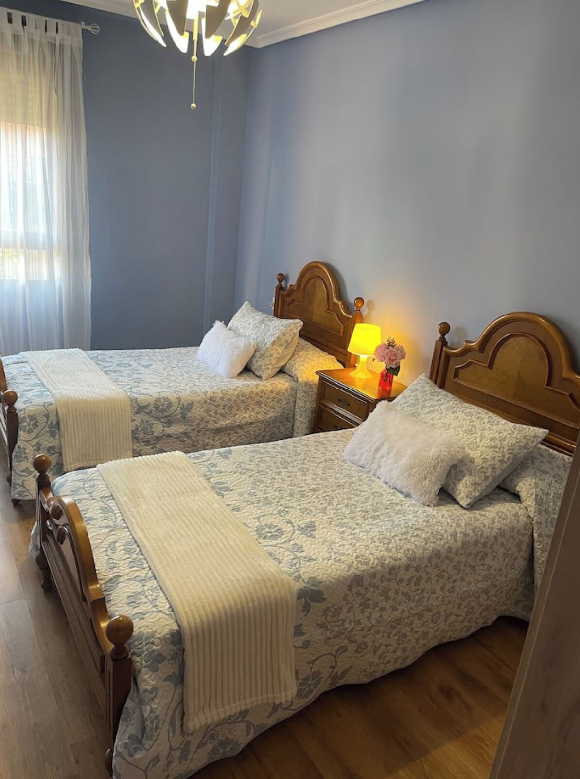 Habitación de hotel con dos camas, cabecero de madera, mesita de noche con lámpara y ventana con cortinas traslúcidas.