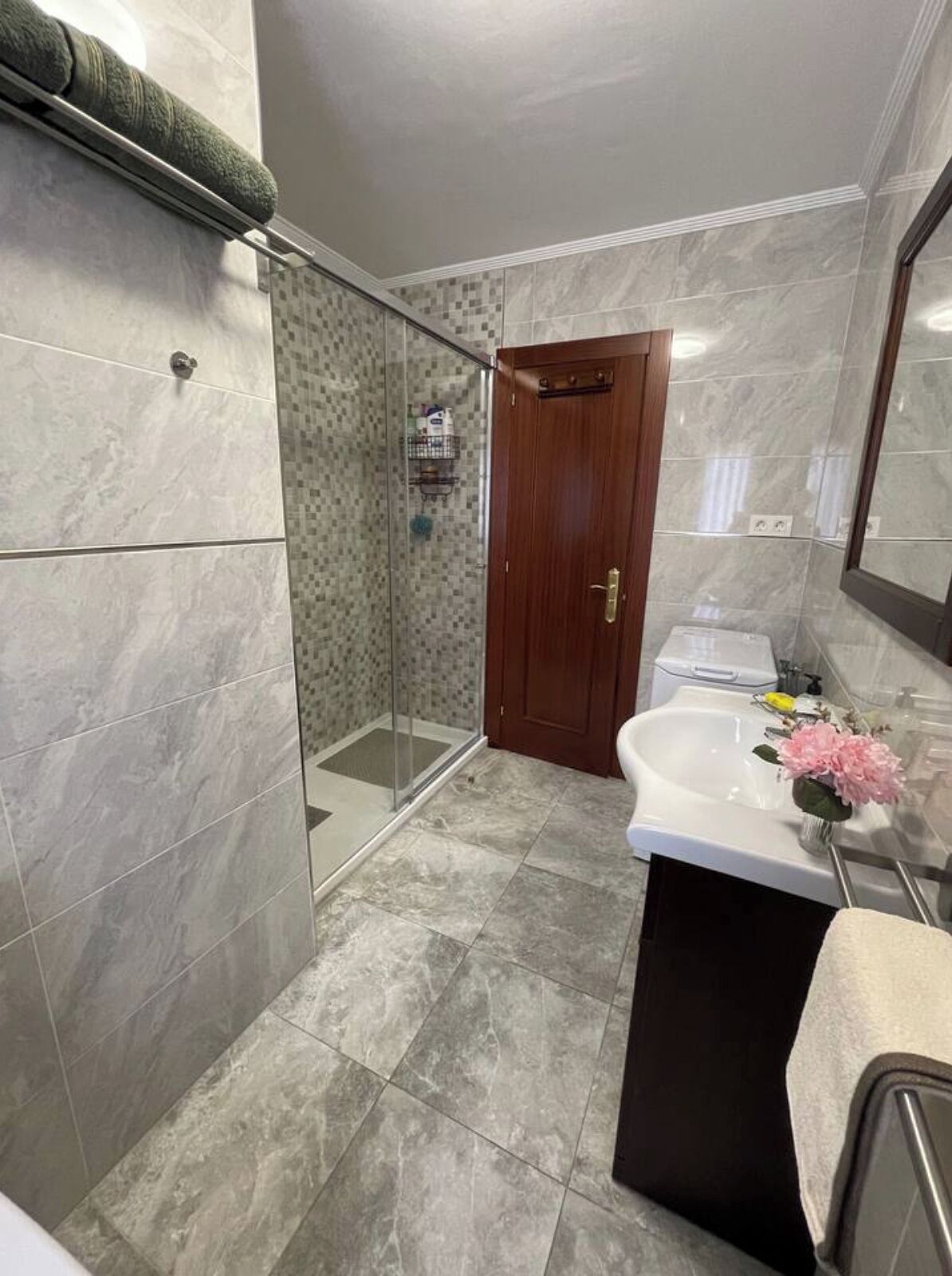 Een moderne badkamer met een glazen douchecabine, een houten deur en een witte wastafel met een bloevas.