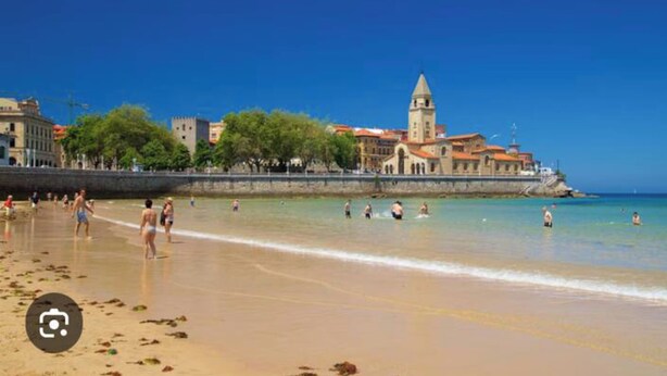 Una playa con gente nadando y caminando por la orilla, cielo despejado y edificios al fondo.