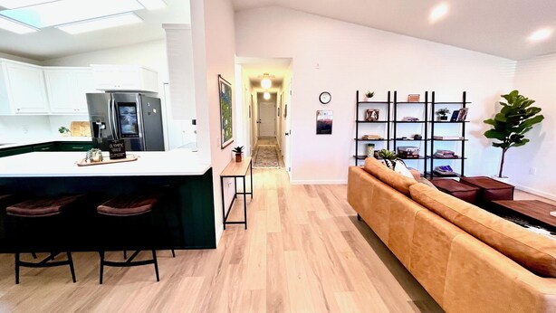 A modern kitchen with white cabinets and a central island, a living area with a sofa and a bookshelf, and a hallway leading to a door. - Olympic House with SAUNA/Rec Room/AC in the Heart of Port Angeles (Port Angeles)