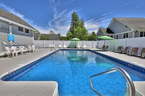 A rectangular swimming pool with a blue tile border, surrounded by white lounge chairs and umbrellas.