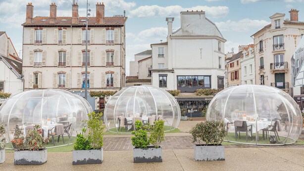Una estructura abovedada de material transparente con mesas y sillas en su interior, rodeada de plantas en macetas.