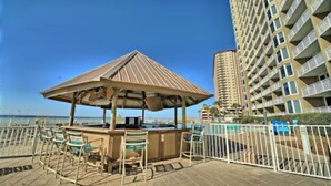 A beachside bar area with a wooden gazebo, chairs, and a pool in the background.