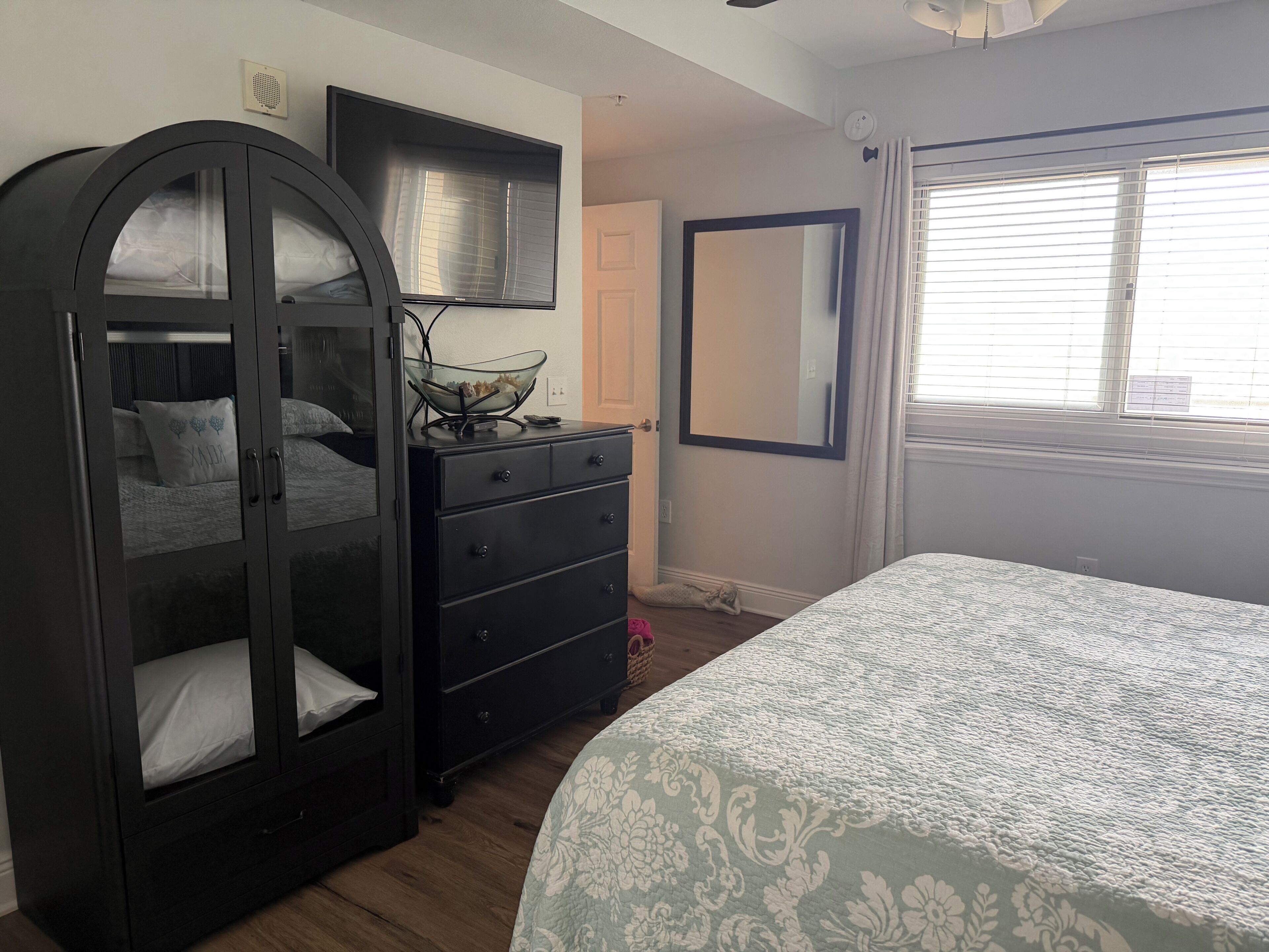 Un dormitorio con cama, cómoda, armario, espejo y ventana con persianas.