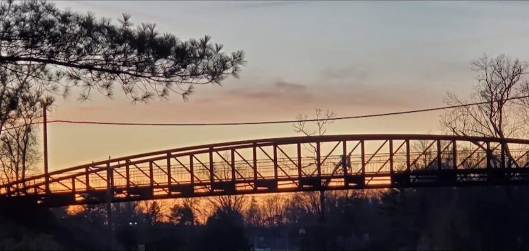 Un puente con estructura de celosía, rodeado de árboles y con un atardecer de fondo.