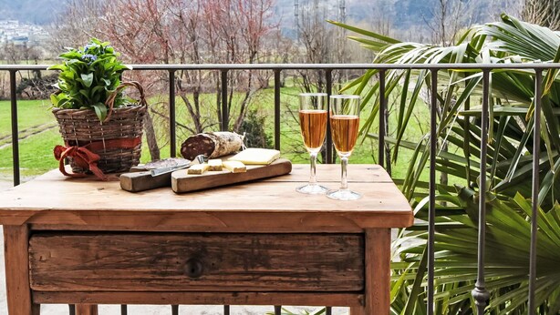 A wooden table with a basket of green plants, a loaf of bread, a cheese platter, and two glasses of wine on a balcony with a metal railing. - Beautiful apartment for 3 people with WIFI, TV, patio and pets allowed (Era)