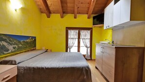 Quarto com cama, teto de madeira, pintura de paisagem de montanhas e área de cozinha com armários e pia.