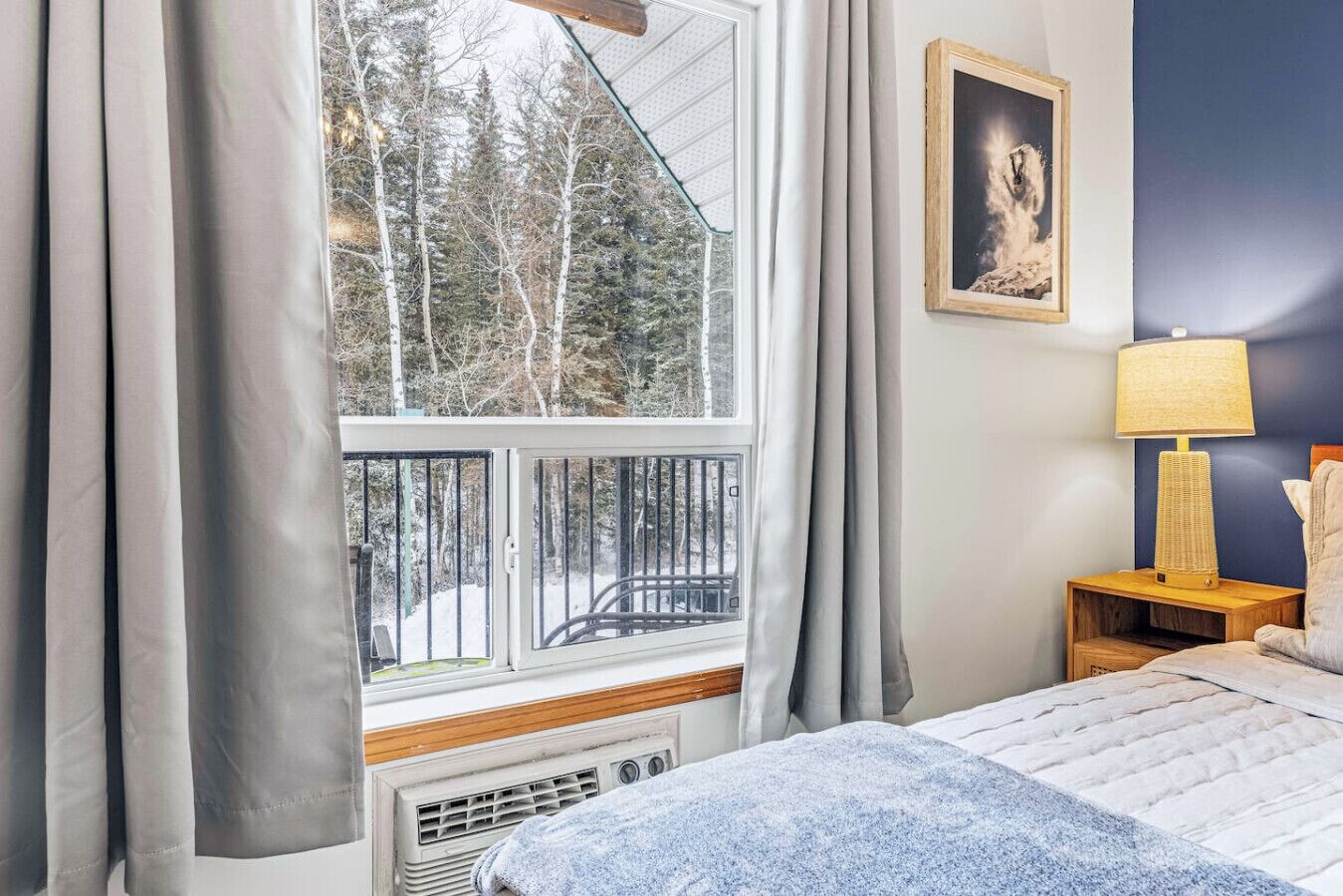 A bedroom with a large window, a bed, a nightstand with a lamp, and a framed picture on the wall.