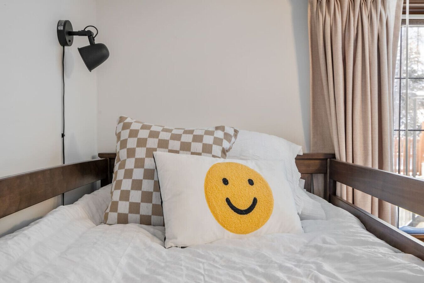Una cama con ropa blanca, una almohada a cuadros, una almohada con cara sonriente, una lámpara de pared y una ventana con cortinas.
