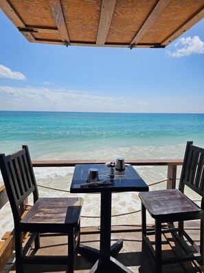 Une table en bois, accompagnée de deux chaises, est placée face à une plage bordée d’une eau turquoise et cristalline.