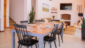 Un coin repas avec une table en bois dressée pour six personnes, entourée de chaises noires. On aperçoit un canapé et un téléviseur à l’arrière-plan.