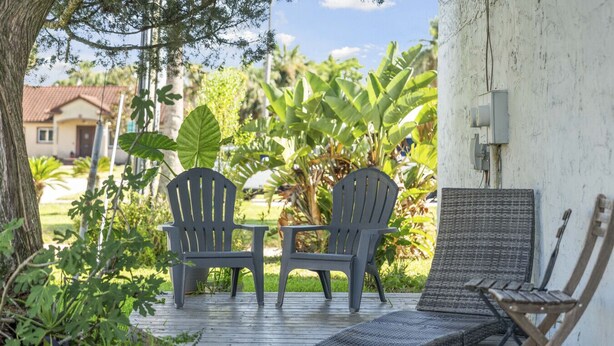 A wooden deck with two Adirondack chairs and a small table, surrounded by lush greenery and a white wall.