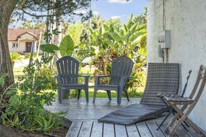 Een houten terras met twee Adirondack-stoelen en een kleine tafel, omringd door weelderige begroeiing en een witte muur.