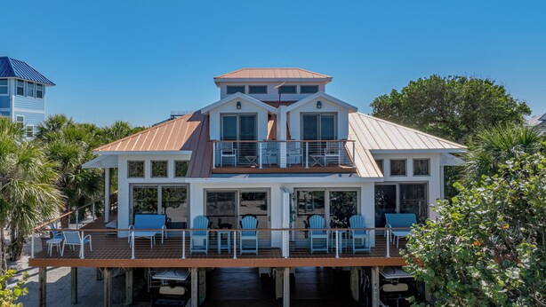 A two-story beachfront house with a covered deck, multiple windows, and a balcony.