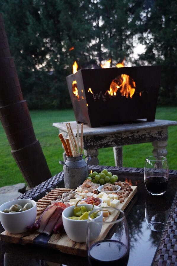 A wooden table with a charcuterie board, wine glasses, and a fire pit in the background.