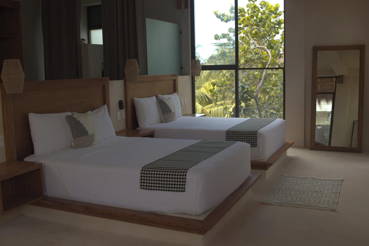 Habitación de hotel con dos camas, un ventanal con vistas a árboles y un espejo en la pared.