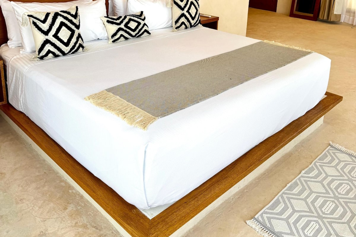 Una cama grande con cabecera de madera, ropa de cama blanca y almohadas estampadas. Hay una mesita de noche pequeña con una lámpara y un espejo. La habitación tiene una alfombra color beige y suelo de madera.