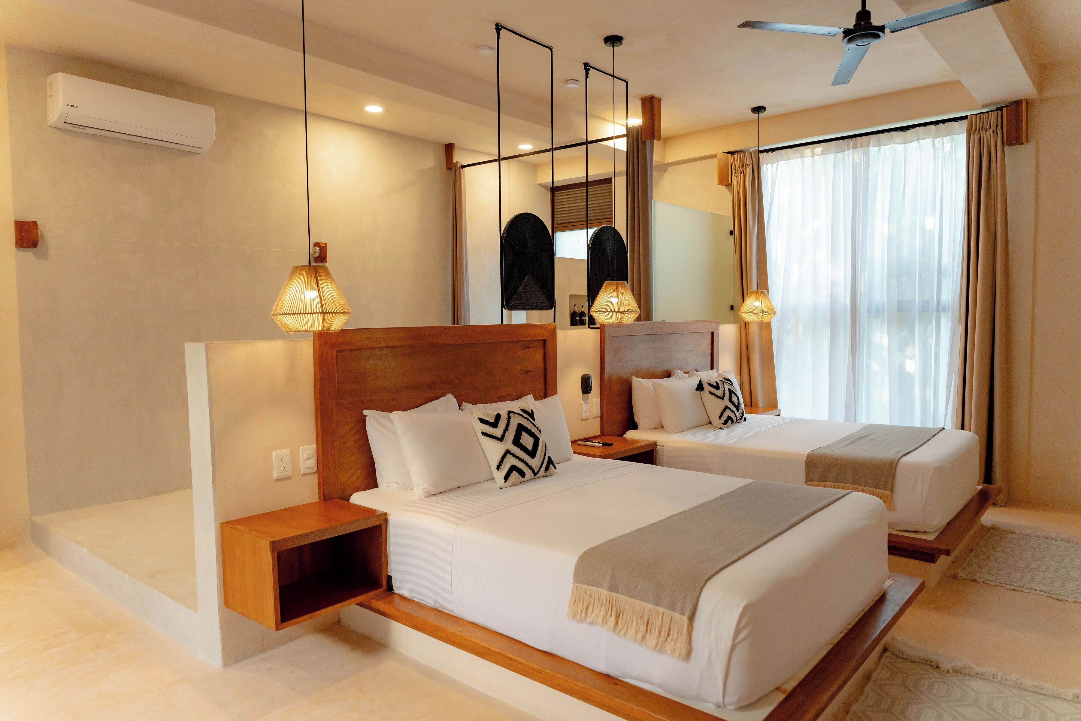 Habitación de hotel con dos camas, ventilador de techo, aire acondicionado y un gran ventanal con cortinas.