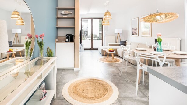 A modern living room with a kitchen, a dining area, and a large mirror. There are wooden shelves, a round woven rug, and a hanging light fixture.