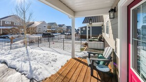 Une véranda couverte avec une terrasse en bois, des sièges d’extérieur et une vue sur une rue résidentielle bordée de maisons et de voitures garées.