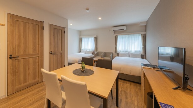 Habitación de hotel con dos camas, un escritorio con computadora, una mesa de comedor con sillas y un televisor.