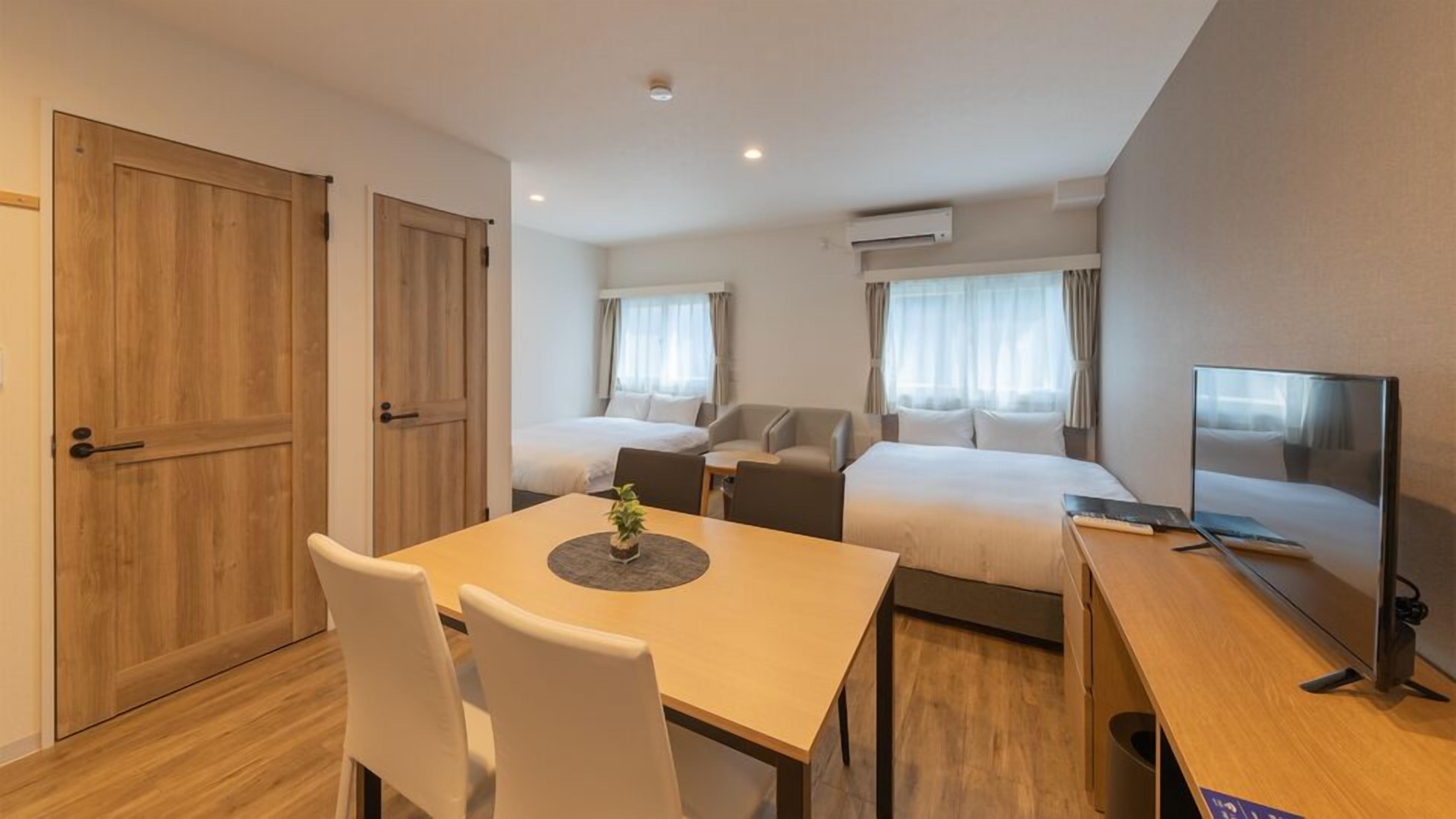 A hotel room with a wooden desk, two single beds, a TV, and a dining table with chairs.