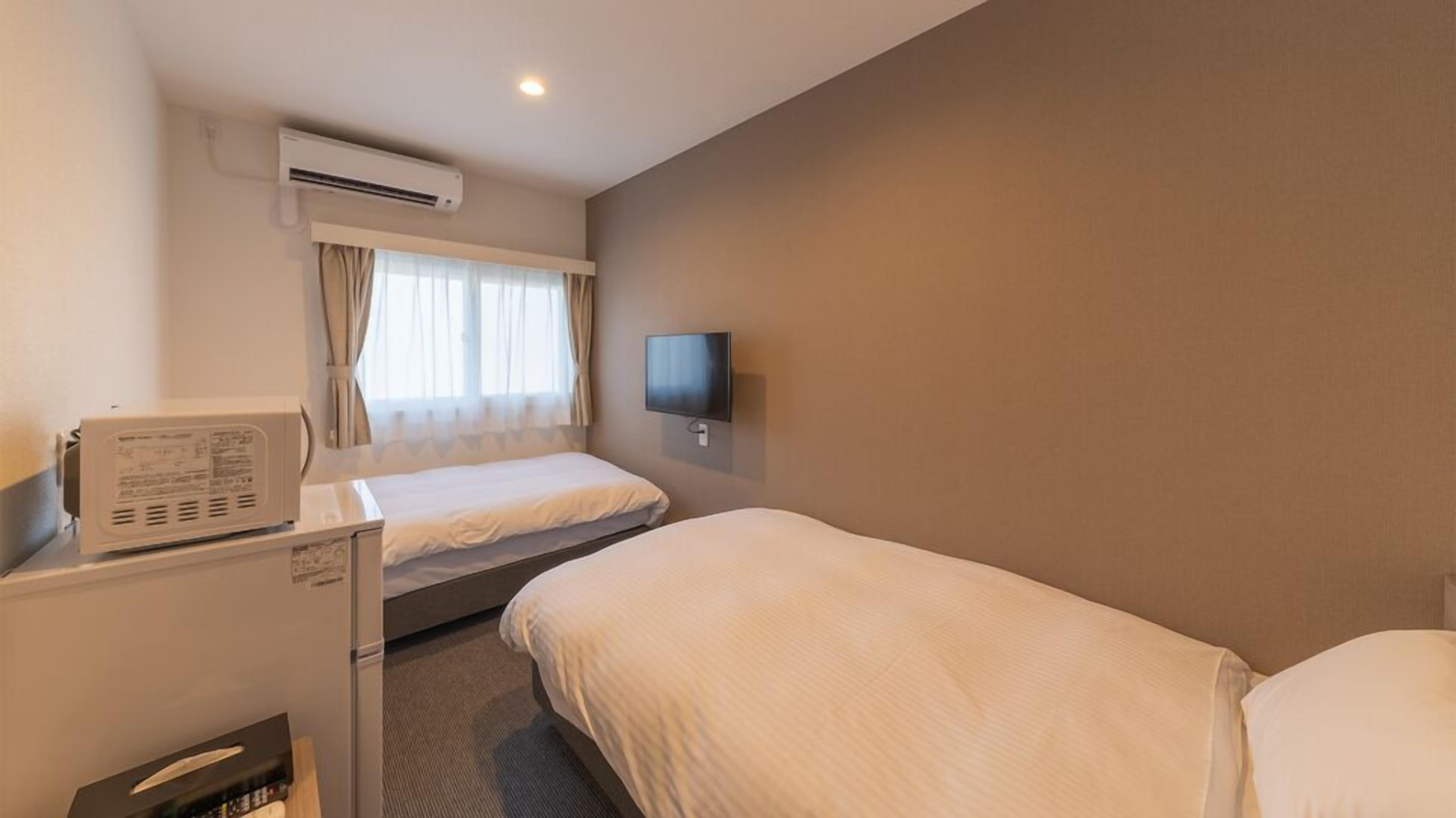 A hotel room with a bed, a desk with a computer, an air conditioner, a TV mounted on the wall, and a window with curtains.