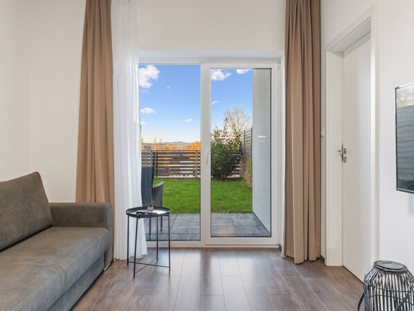 A modern living room with a grey sofa, a small round table, and a sliding glass door leading to a garden.