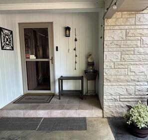 A front porch with a door, a bench, a wall-mounted light fixture, a small table, and a potted plant.