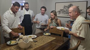 A group of people dining at a wooden table with a chandelier above, in a room with a white ceiling and a framed picture on the wall.